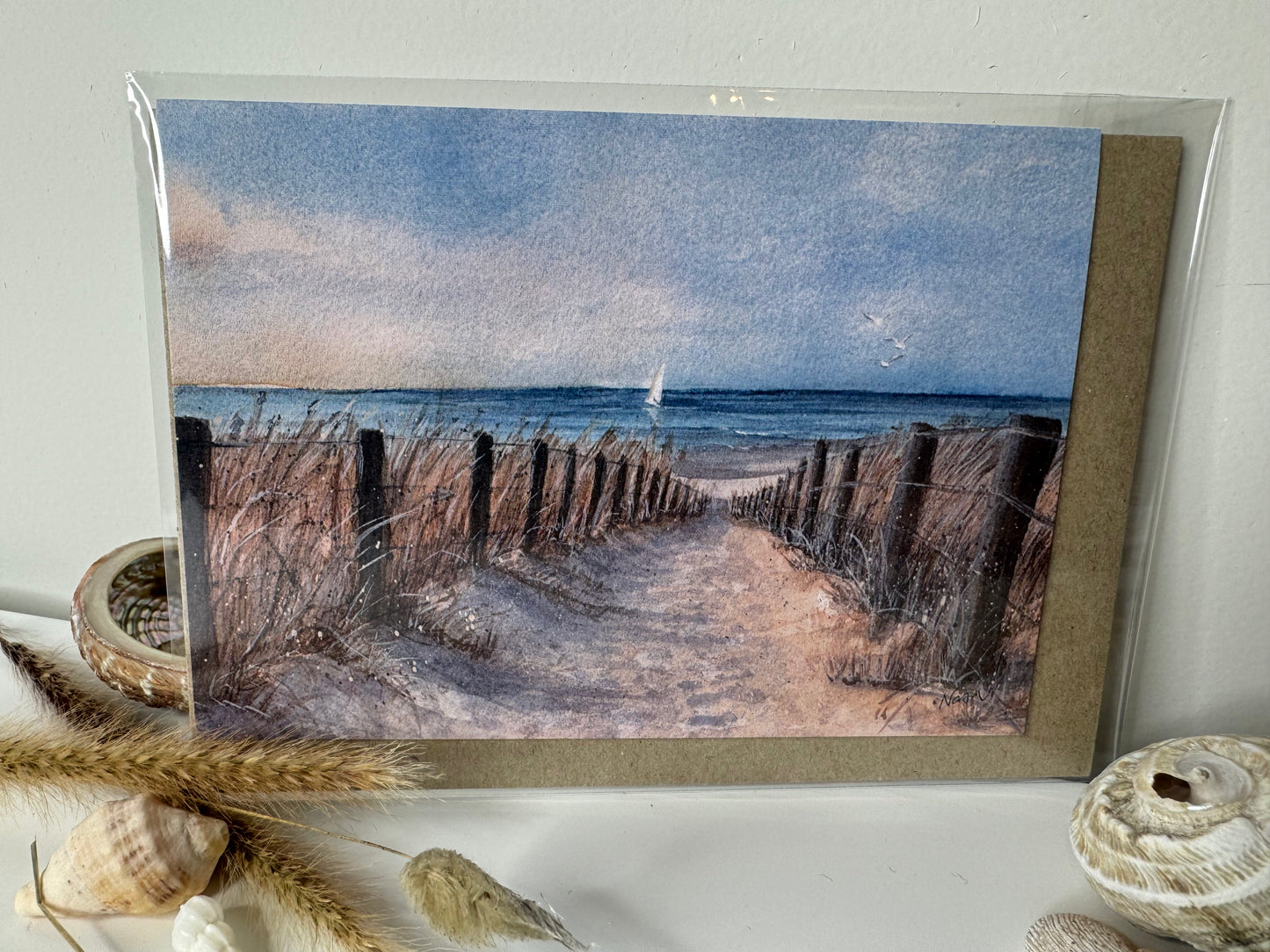 Image of actual card in cellophane envelope for shipping. Image depicts a sandy path leading down to a beach with sailboat in the distance.